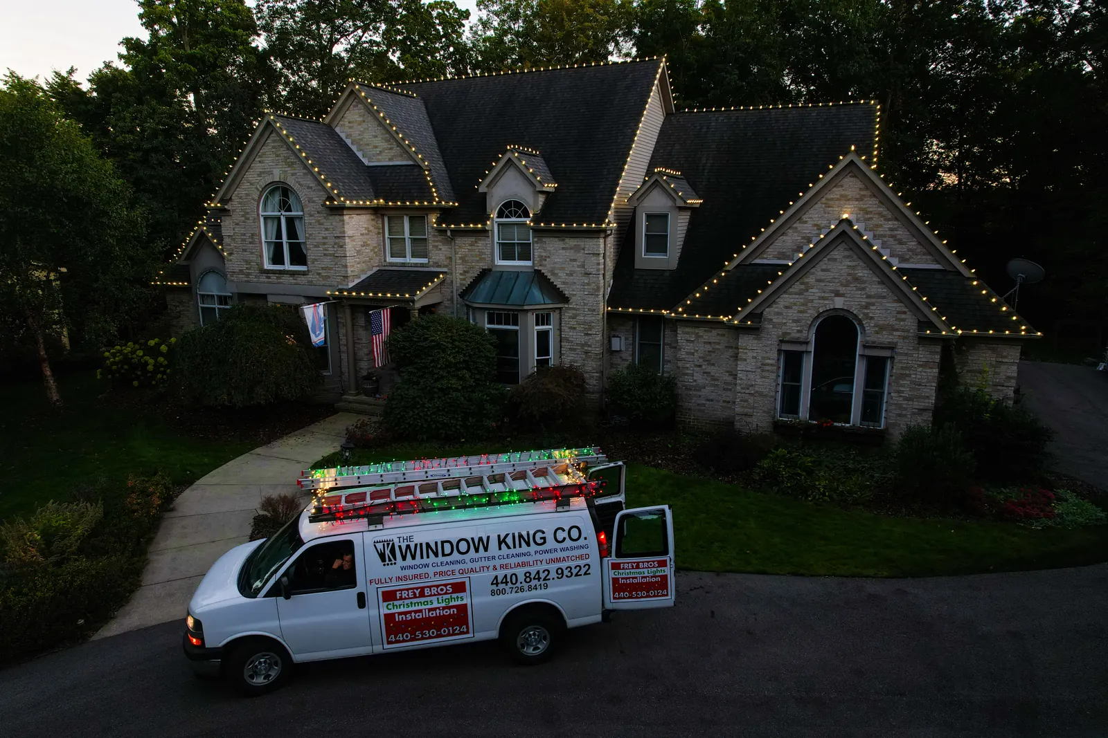 Frey Bros Christmas lights van parked in a driveway at dusk in front of a stone home with warm white lights on every gable