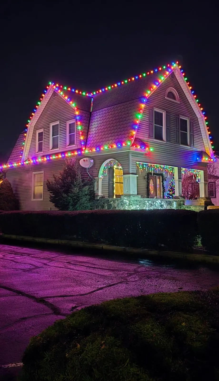Dutch Colonial home at night with multicolor C9 Christmas lights on both gambrel roofs, icicle lights on the porch, and a lit stone porch base
